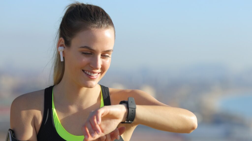 Woman wearing wireless earbuds and a smart watch checks her wrist while smiling during an outdoor workout.